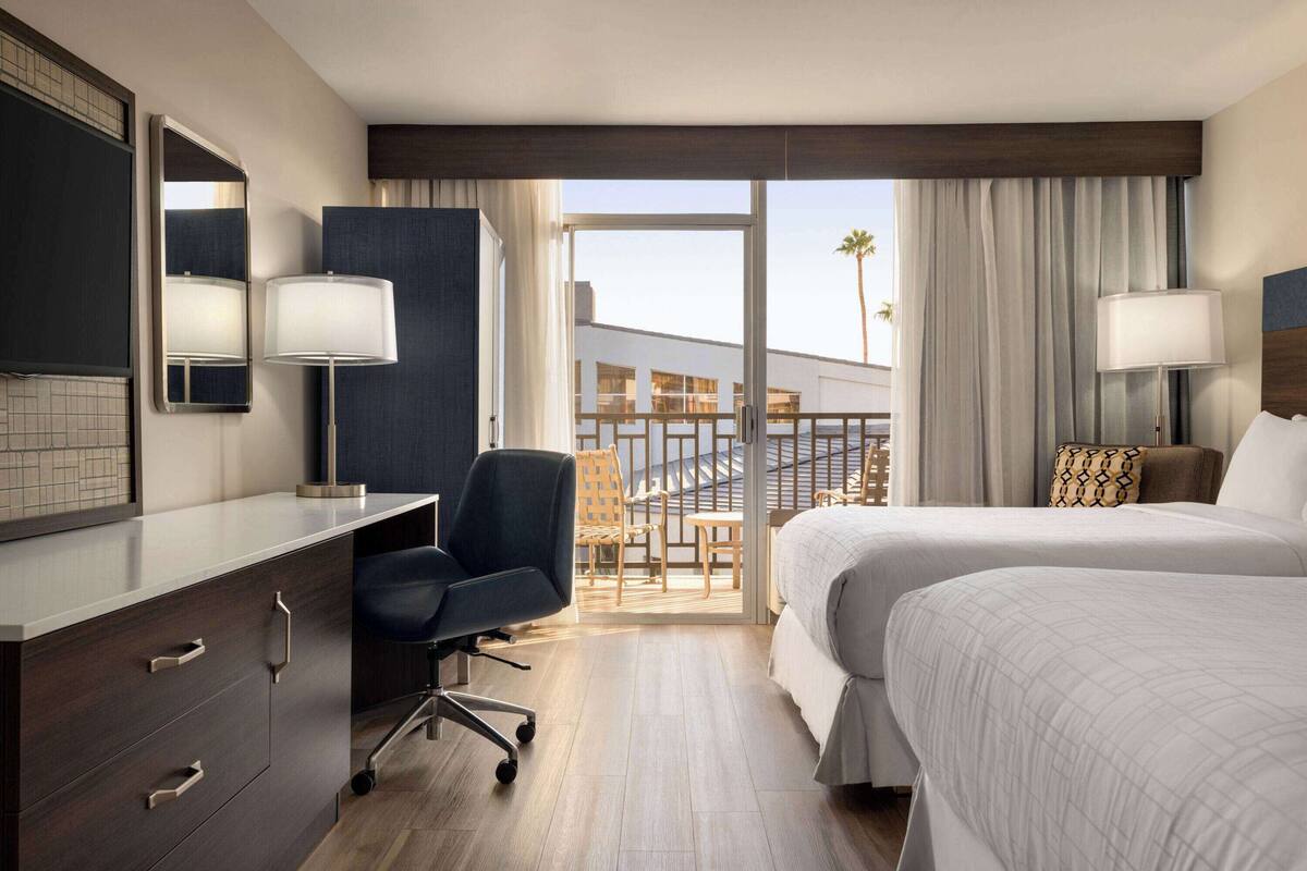 A hotel room with a desk, two beds, a chair, and a balcony with a view of palm trees.