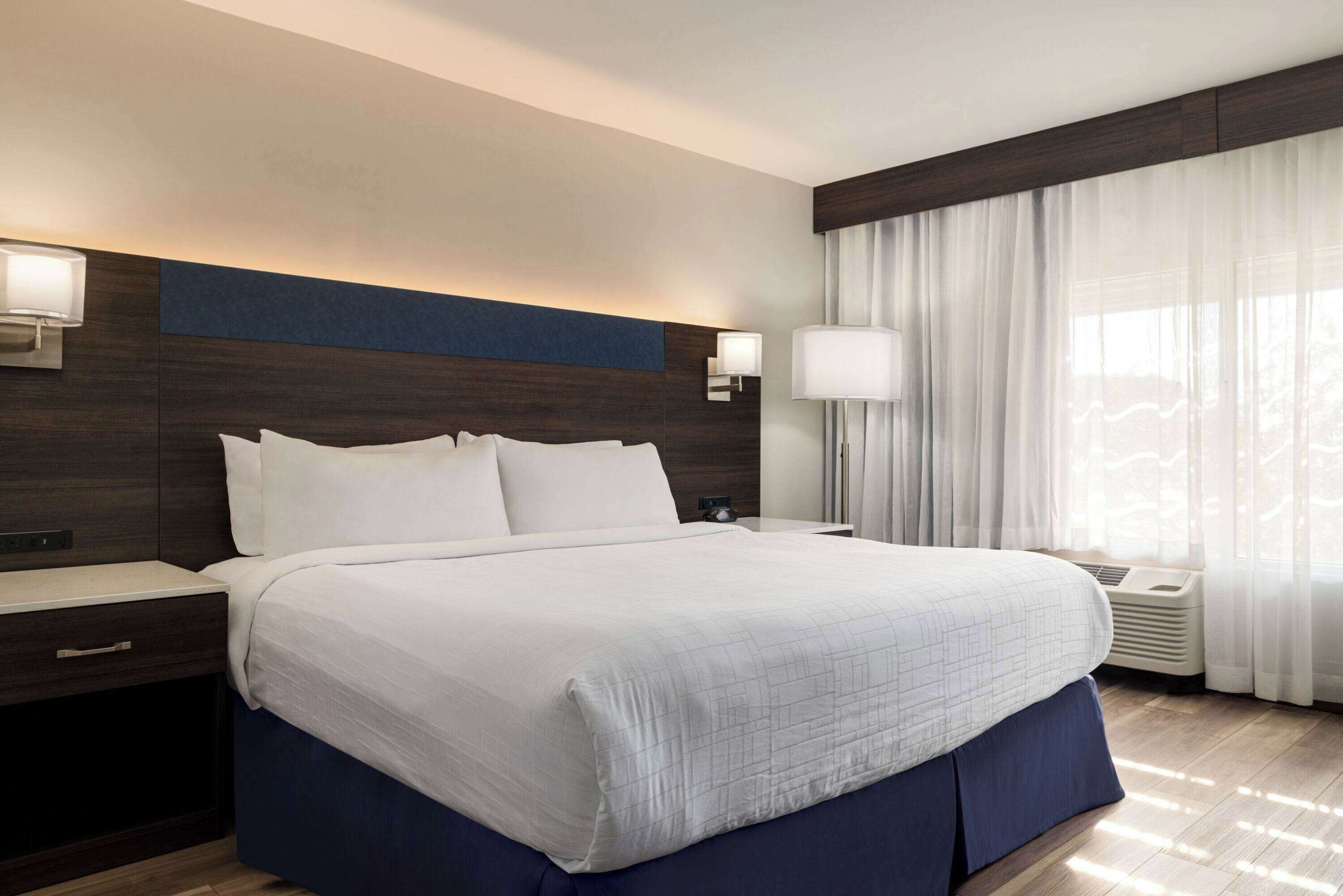 Ett modernt hotellrum med en stor säng, ett sängbord, en liten avlastningsyta, en vägglampa och ett fönster med tunna, genomskinliga gardiner.