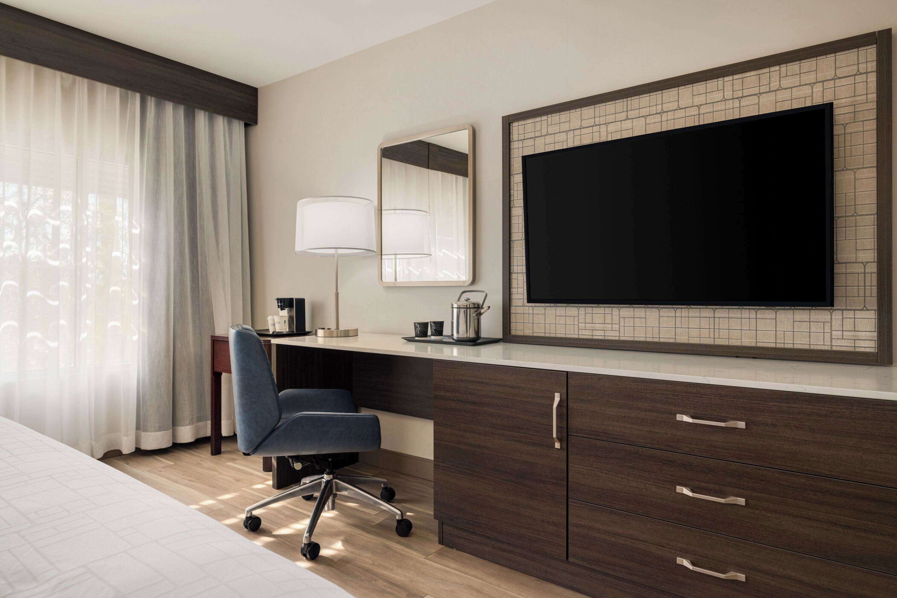 A hotel room with a bed, a desk with a chair, a flat-screen TV, a lamp, and a mirror.