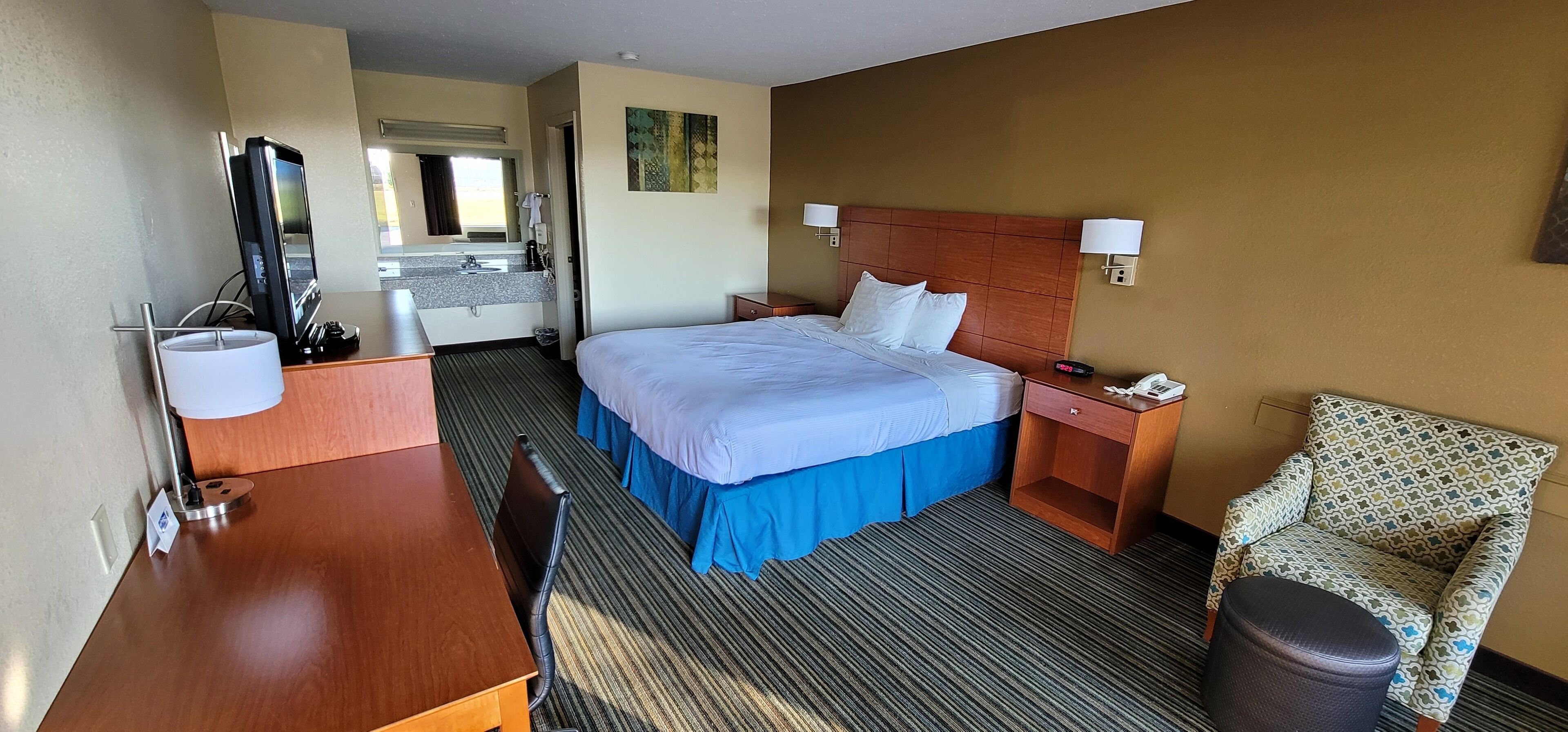 Une chambre d’hôtel avec un lit, un bureau, une chaise, un miroir et une salle de bain visible à travers une porte ouverte.
