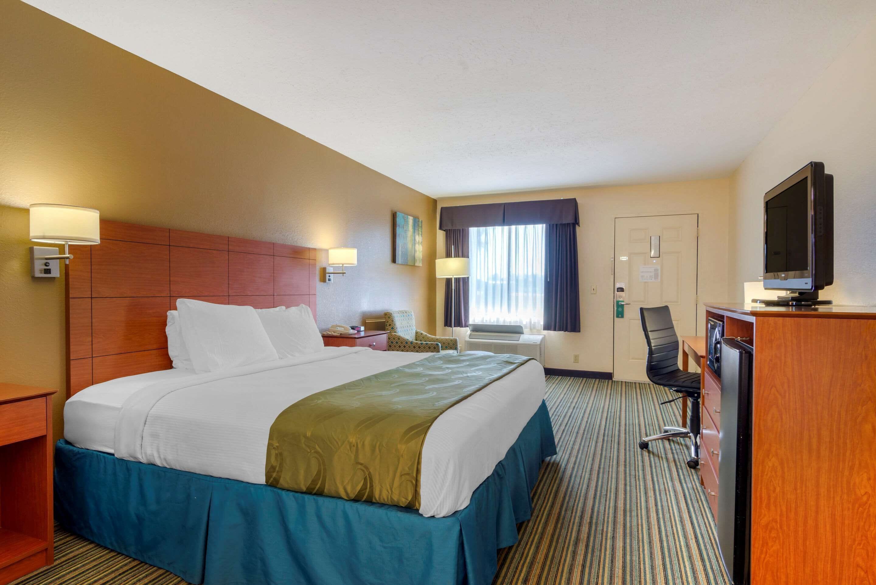 A hotel room with a large bed, a desk with a computer, a chair, a television, and a window with curtains.
