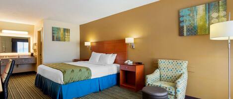 A hotel room with a bed, a chair, a desk, a trash can, and a painting on the wall.