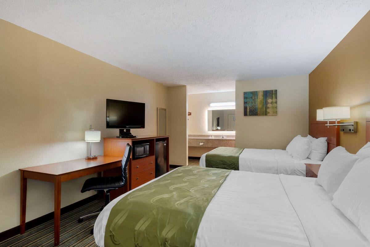 Quarto de hotel com duas camas, uma escrivaninha com cadeira, televisão e banheiro ao fundo.