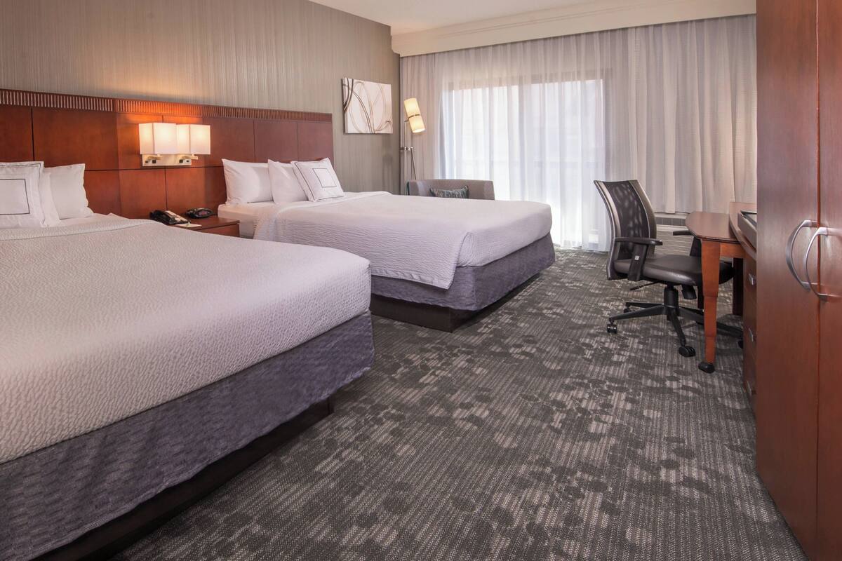 A hotel room with two beds, a desk, a chair, a lamp, and a large window with curtains.