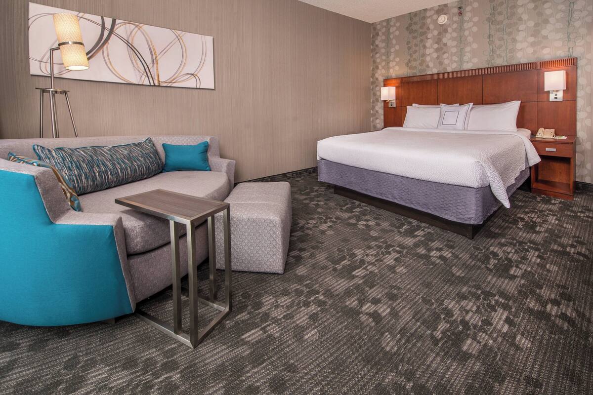 A hotel room with a bed, a sofa, a small table, and a nightstand with a lamp.