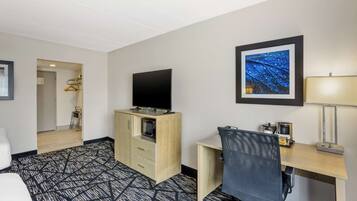 Una habitación de hotel con una cama, un escritorio con una silla, un televisor, un cuadro enmarcado en la pared y suelo alfombrado.