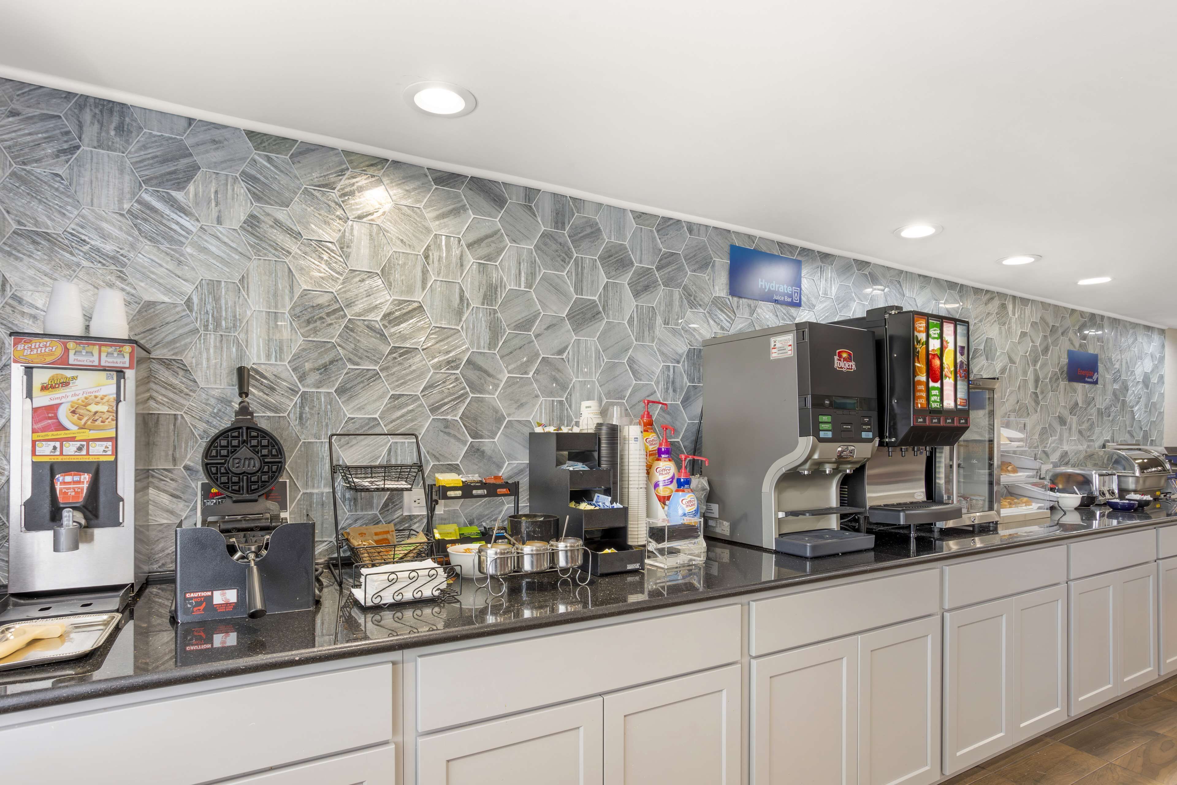 Zona de bufé con diversas estaciones de comida, que incluyen una máquina para gofres, máquinas de café y un mostrador refrigerado.