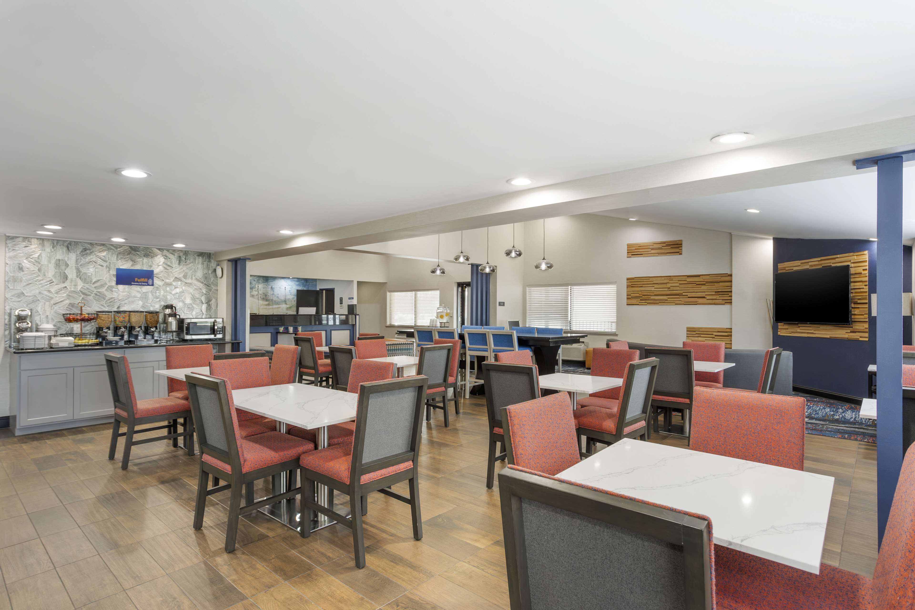 A modern dining area with tables and chairs, a kitchen in the background, and a flat-screen TV mounted on the wall.