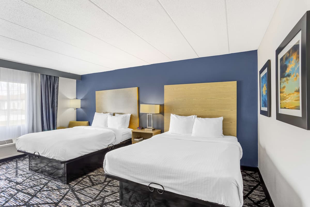 A hotel room with two beds, a large window with curtains, a desk with a lamp, and framed artwork on the wall.