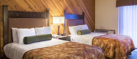 A hotel room with two beds, wooden paneling on the walls, a nightstand with a lamp, and a window with curtains.
