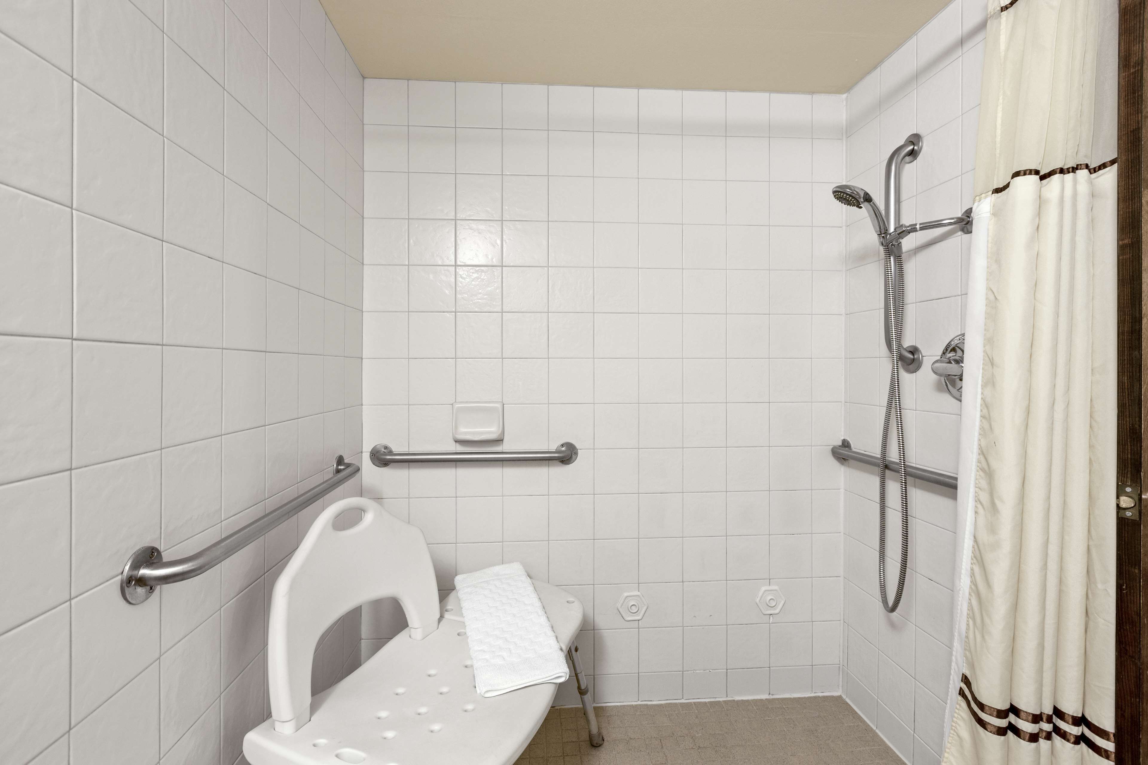 A bathroom with white tiles, a shower with a handheld showerhead, a grab bar, and a white toilet.