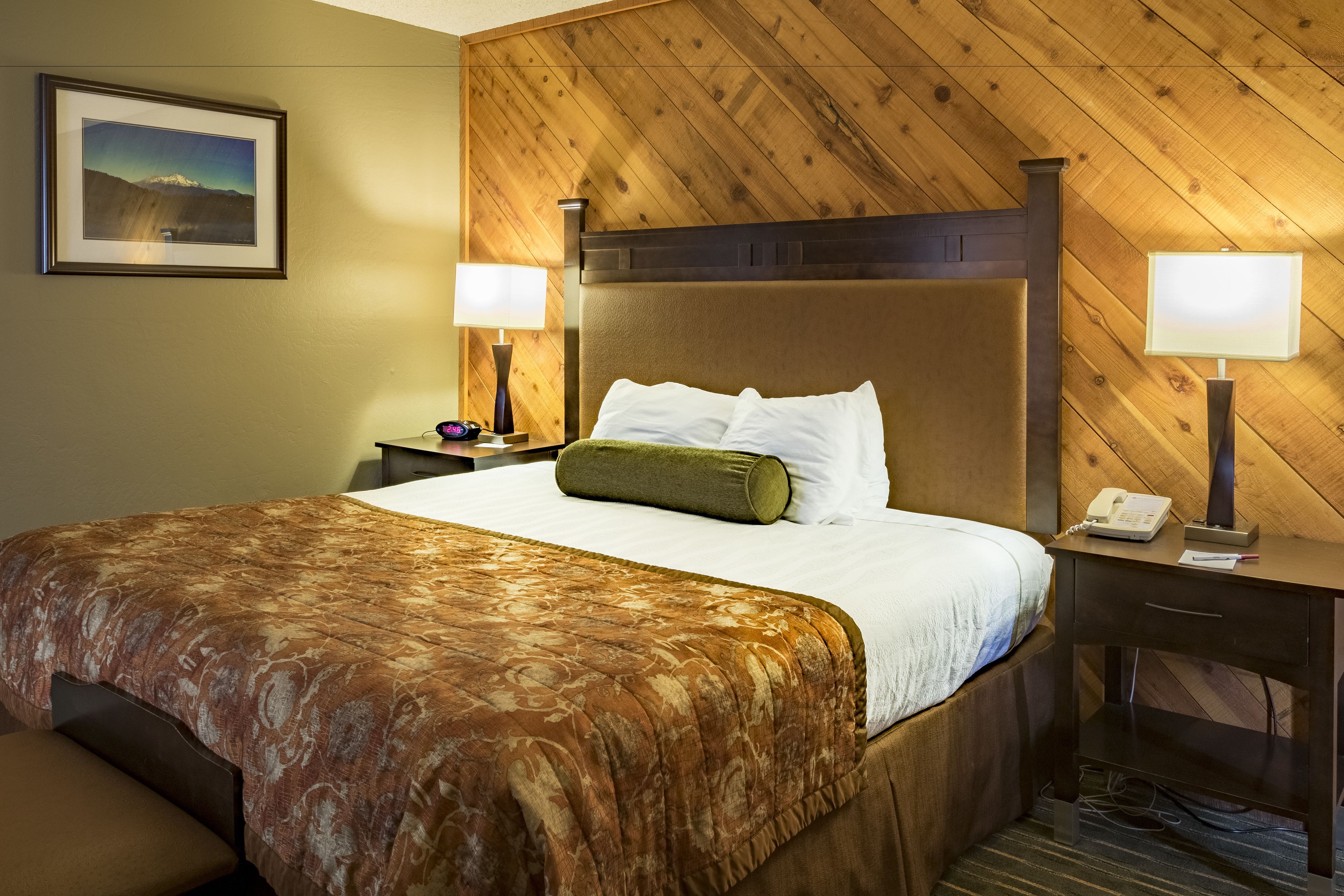 Une chambre d’hôtel avec un grand lit, une tête de lit en bois, deux tables de chevet avec des lampes, un tableau encadré au mur et un téléphone sur l’une des tables.
