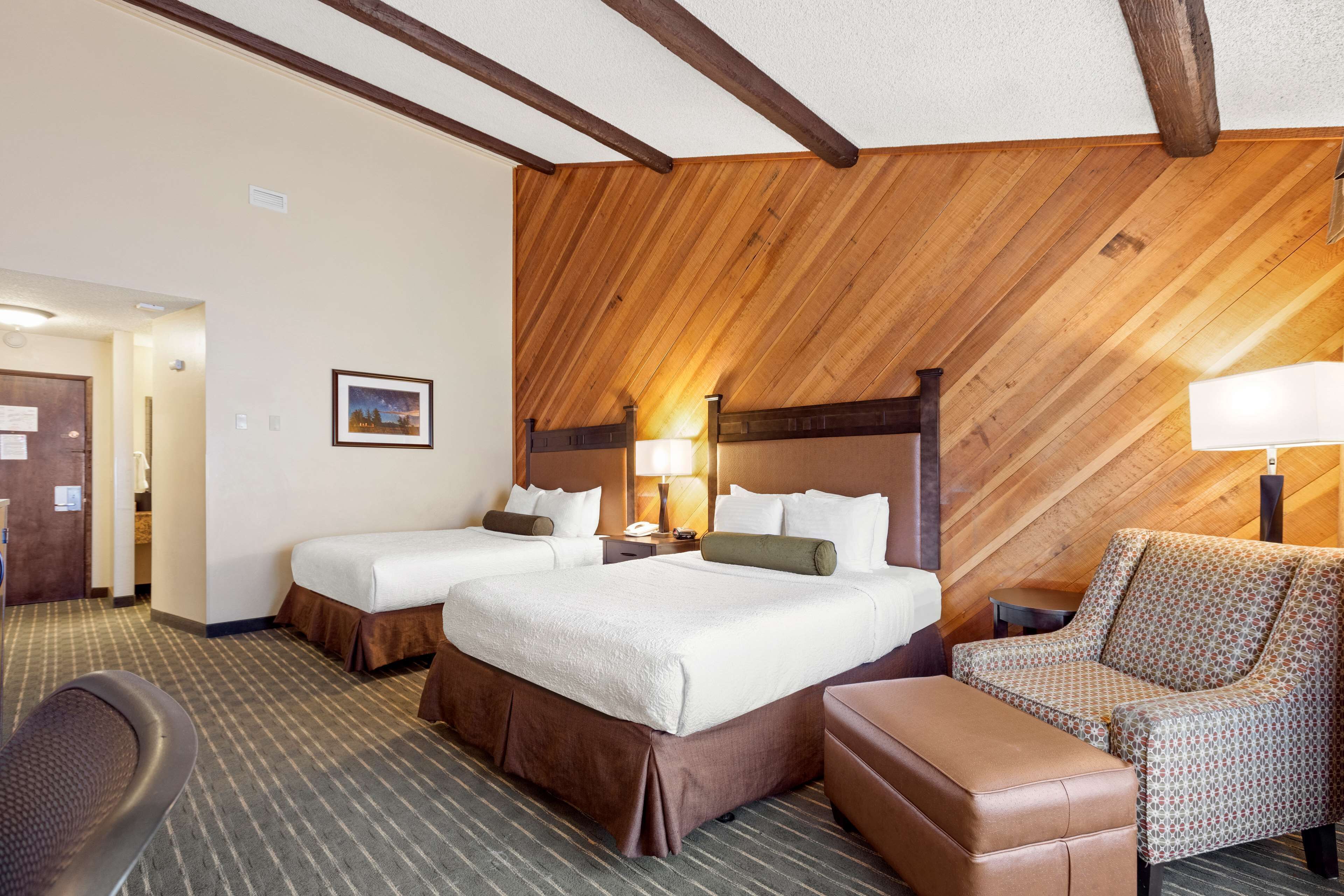 A hotel room with two beds, a wooden ceiling, a patterned armchair, and a small table.