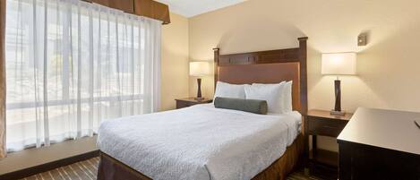 A hotel room with a large bed, two bedside tables with lamps, a desk, and a window with sheer curtains.