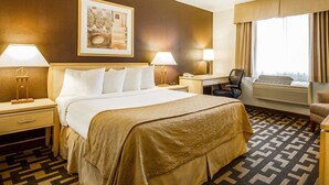 Ein Hotelzimmer mit einem großen Bett, zwei Nachttischen mit Lampen, einem Schreibtisch mit Stuhl, einem Fenster mit Vorhängen und einem an der Wand hängenden Gemälde.