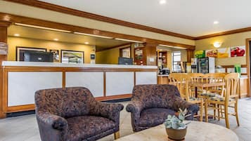 A hotel lobby with a reception desk, two armchairs, a coffee table with a plant, and a breakfast area with wooden chairs and tables.