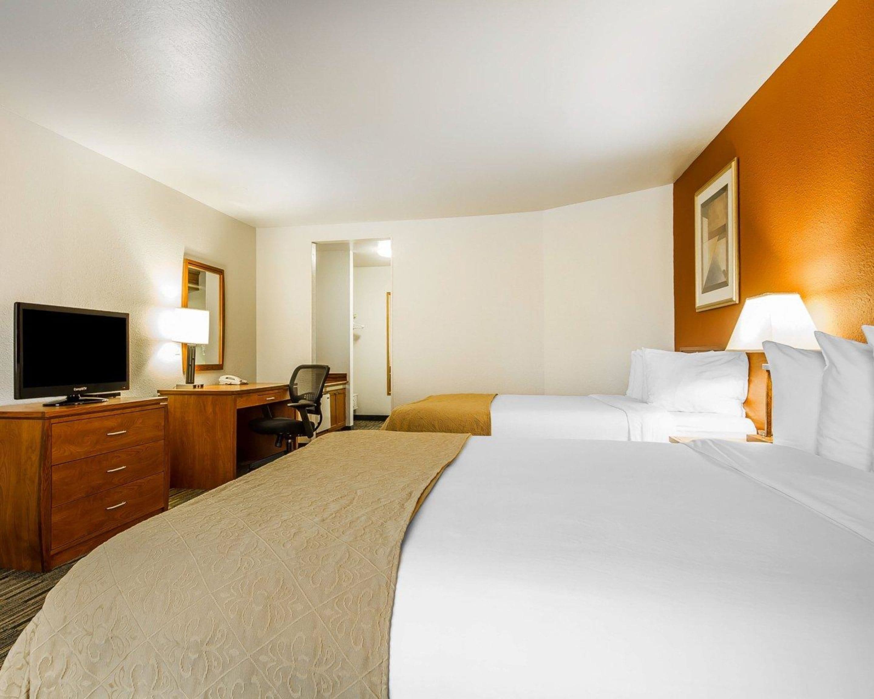 A hotel room with a large bed, a desk with a television, a chair, and a desk lamp.