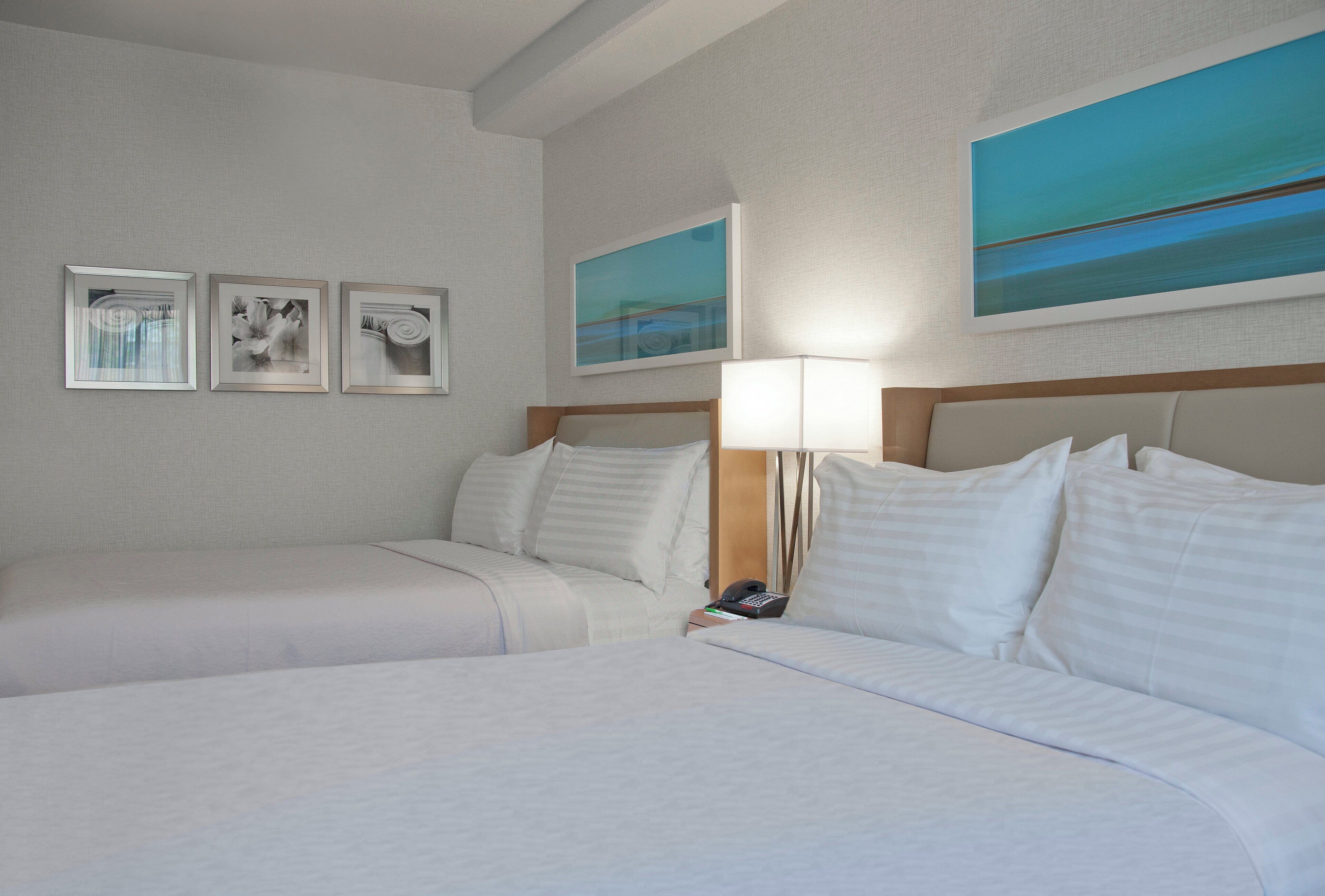 Habitación de hotel con dos camas, una mesita de noche con lámpara y tres cuadros enmarcados en la pared.