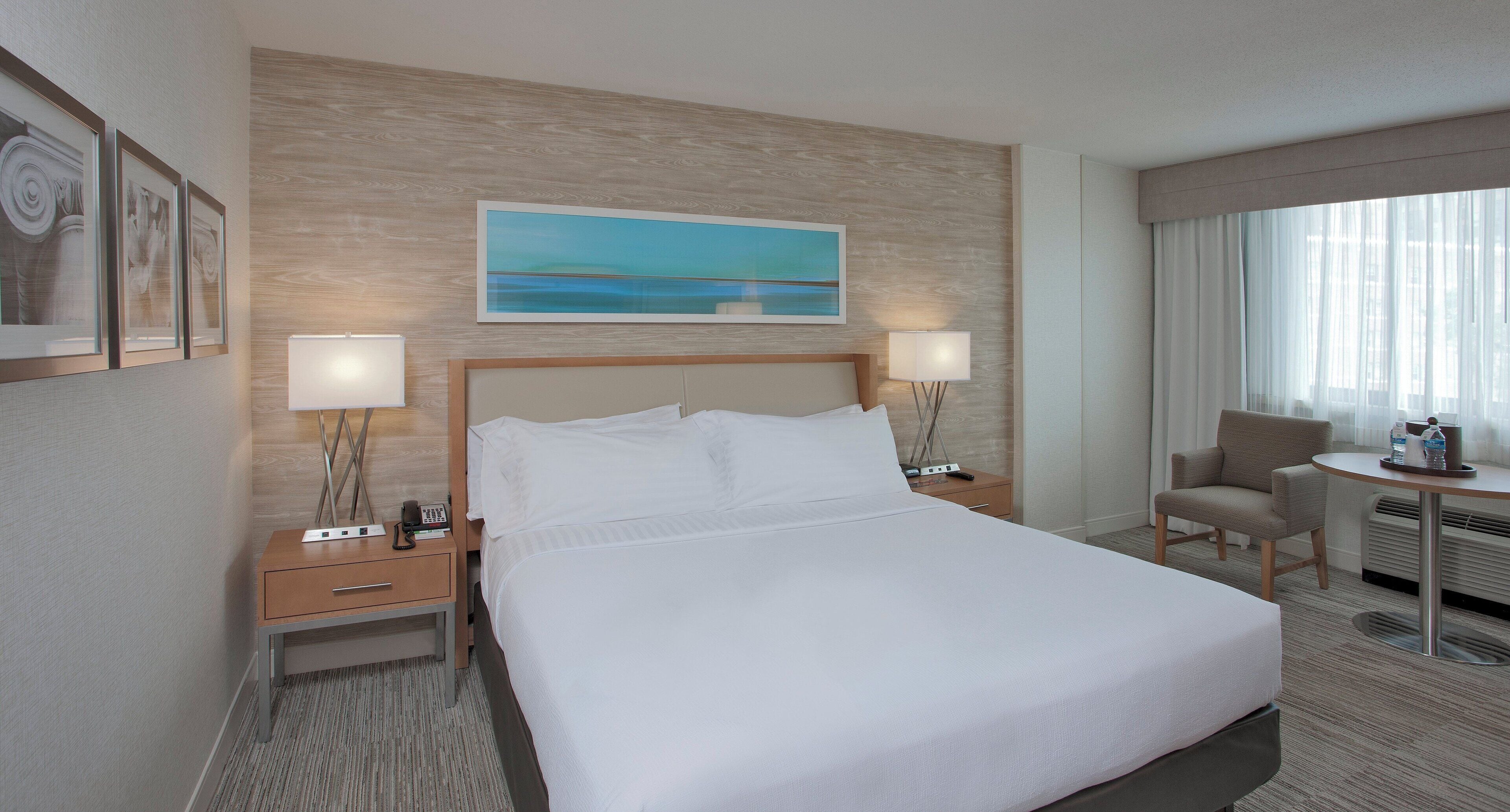 A hotel room with a large bed, two bedside tables with lamps, a desk with a chair, a painting on the wall, and a view of the outdoors through the window.