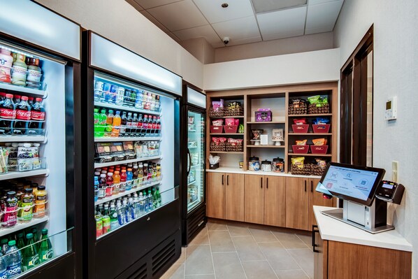 A self-service kiosk area with a touchscreen display, refrigerated beverage dispensers, and shelves stocked with snacks and beverages.