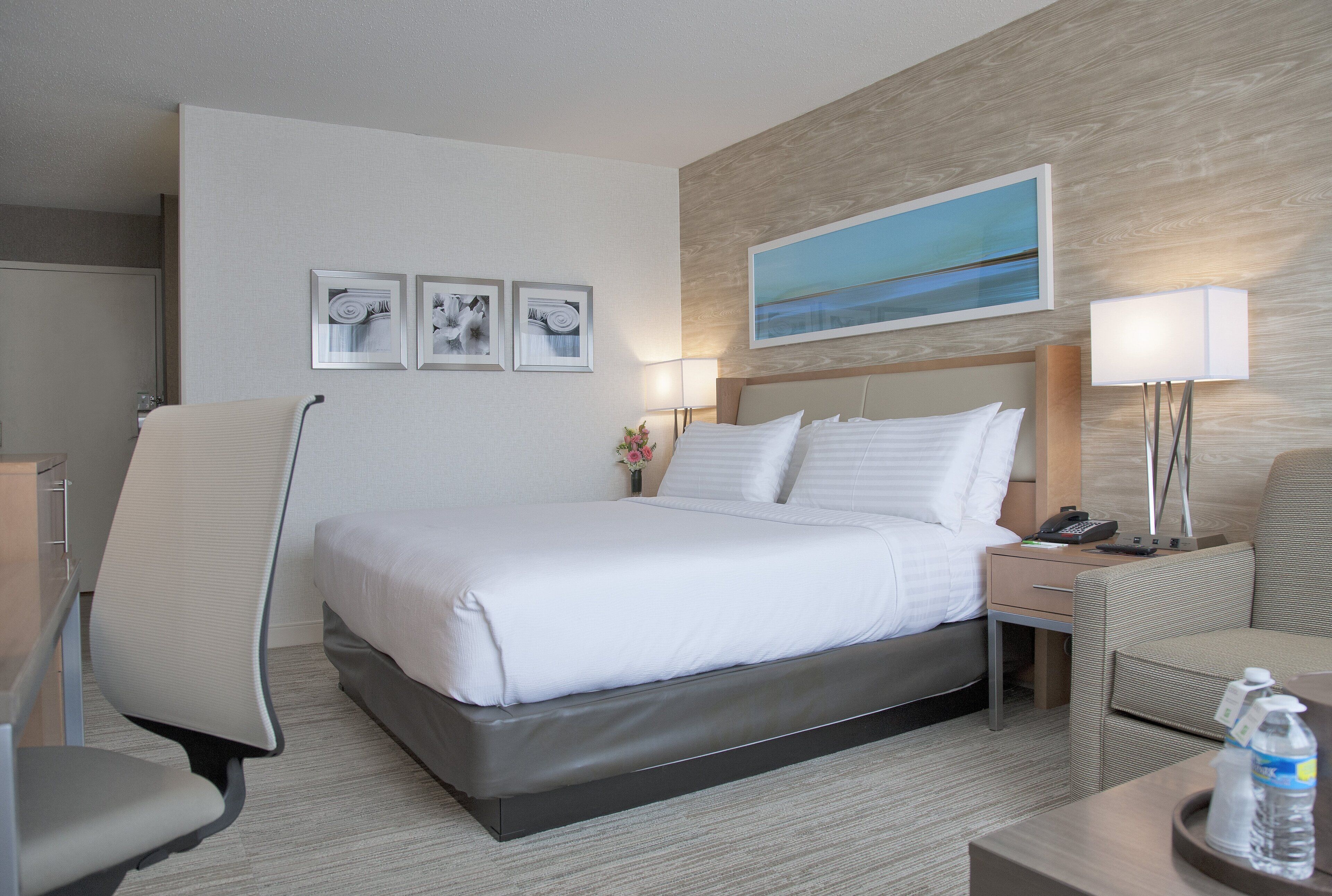 A hotel room with a large bed, a chair, a desk, and a nightstand with a lamp.