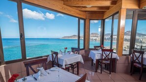 Un restaurante con mesas dispuestas para comer, grandes ventanales que ofrecen vista al mar y un horizonte urbano distante.