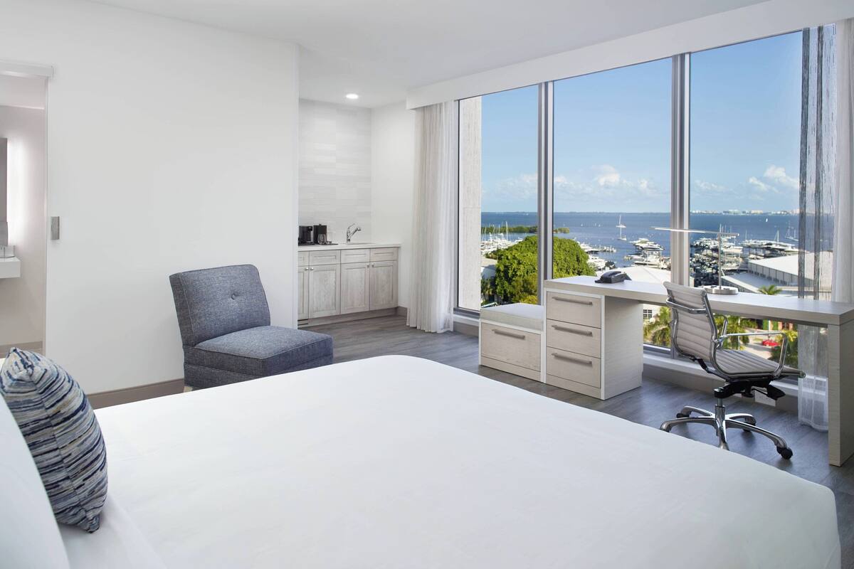 A modern hotel room with a large bed, a desk with a view, a chair, and a small kitchenette.