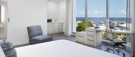 A modern hotel room with a large bed, a desk with a view, a chair, and a small kitchenette.