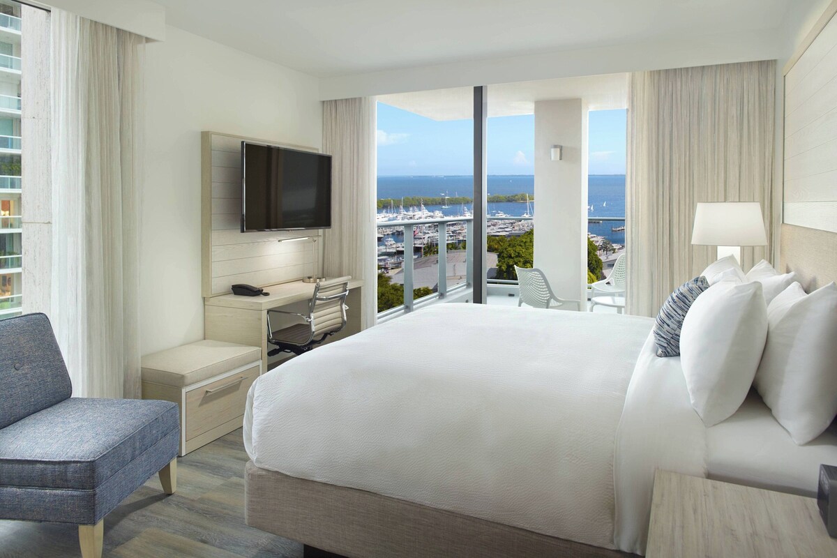 A hotel room with a large bed, a desk with a flat-screen TV, a chair, and a view of the marina and ocean.