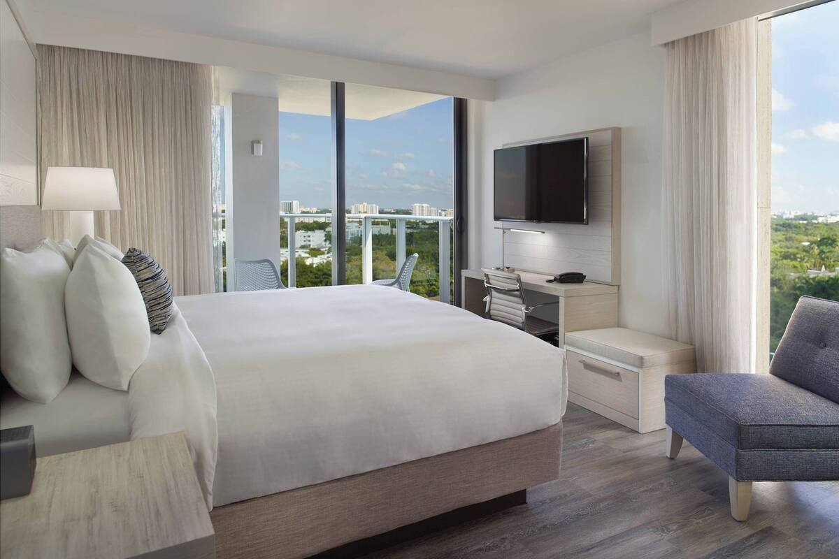 A modern hotel room with a large bed, a desk with a chair, a flat-screen TV, and a city view.