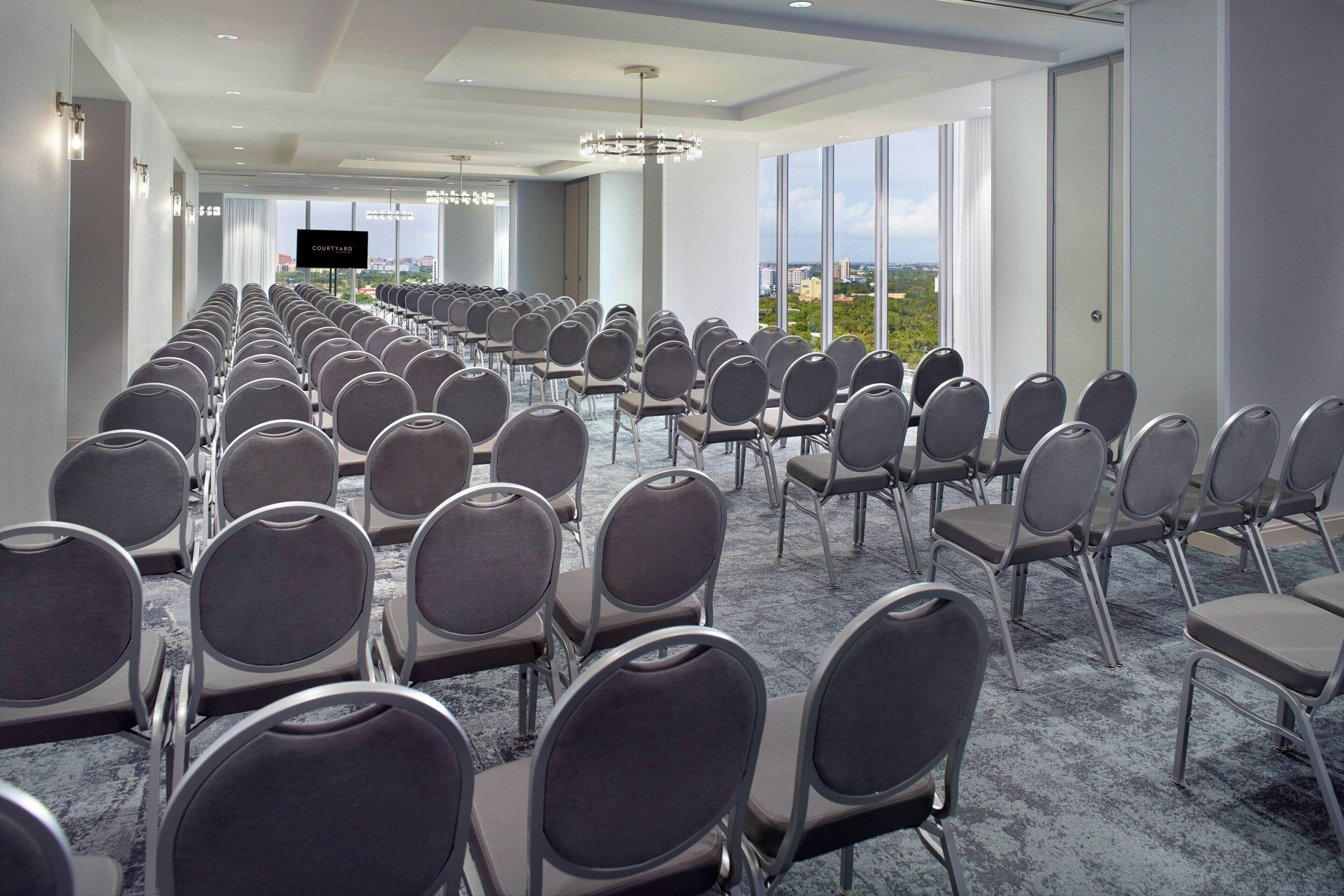Een congreszaal met rijen stoelen, een groot scherm en een uitzicht op de stad door grote ramen.