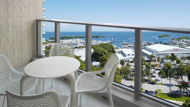 A balcony with a white table and chairs, offering a view of a marina with boats and a large building in the distance.