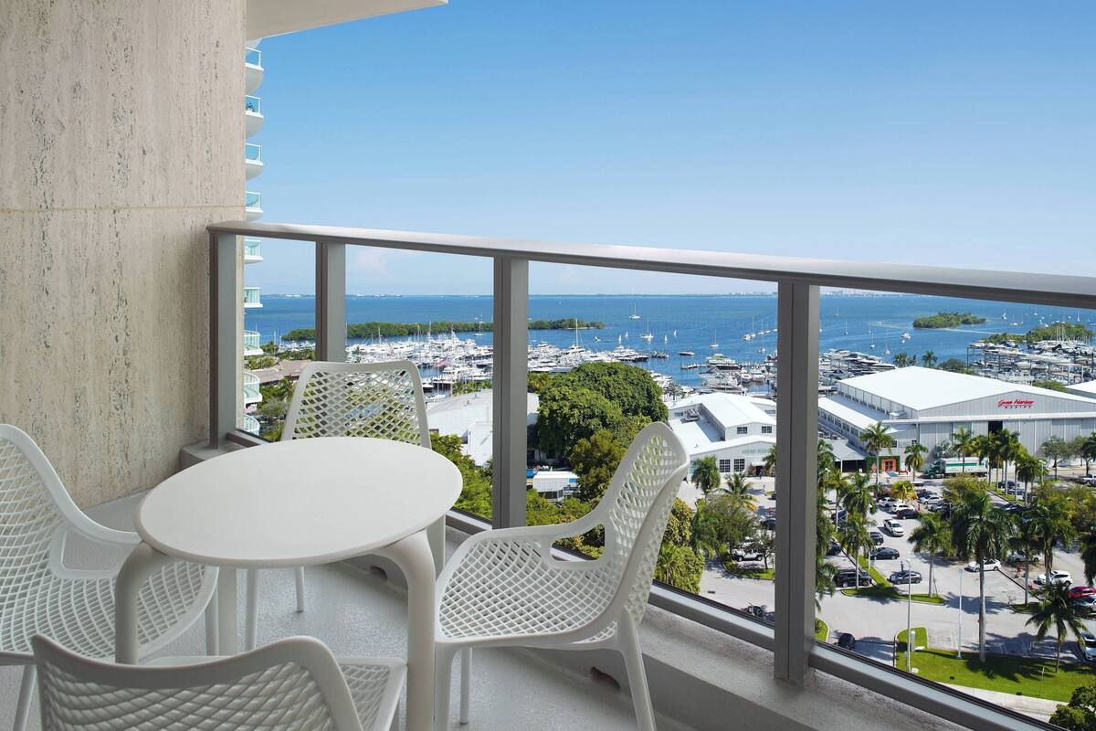 A balcony with a white table and chairs, offering a view of a marina with boats and a large building in the distance.