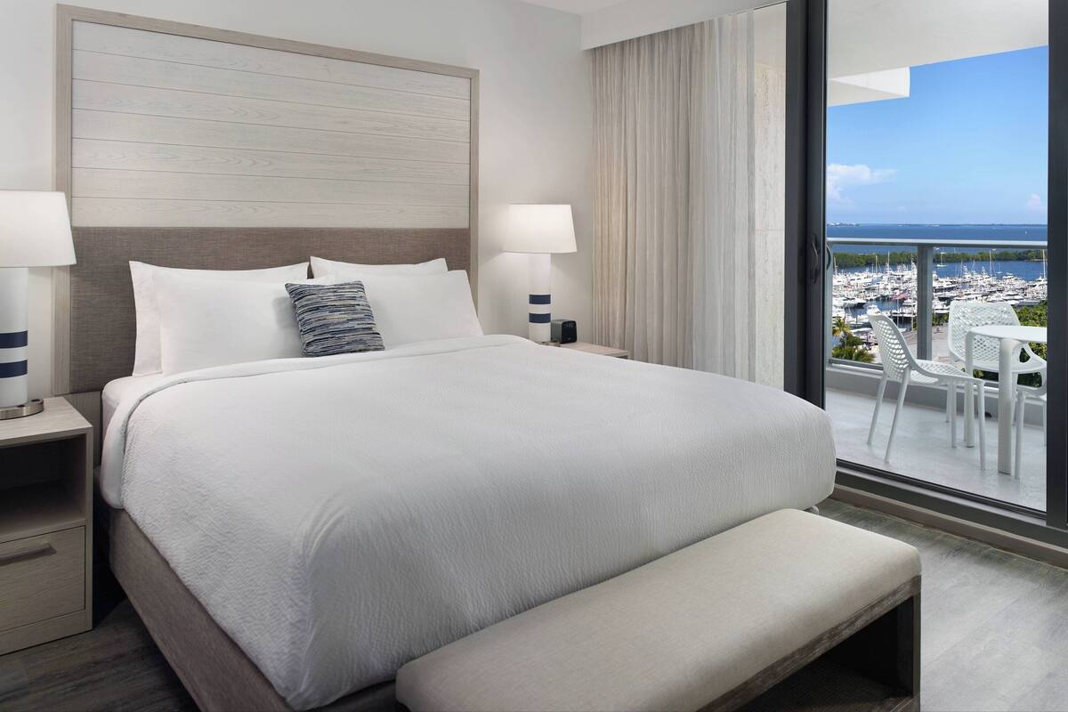 A hotel room with a large bed, two bedside tables, a bench, and a balcony with a view of a marina.