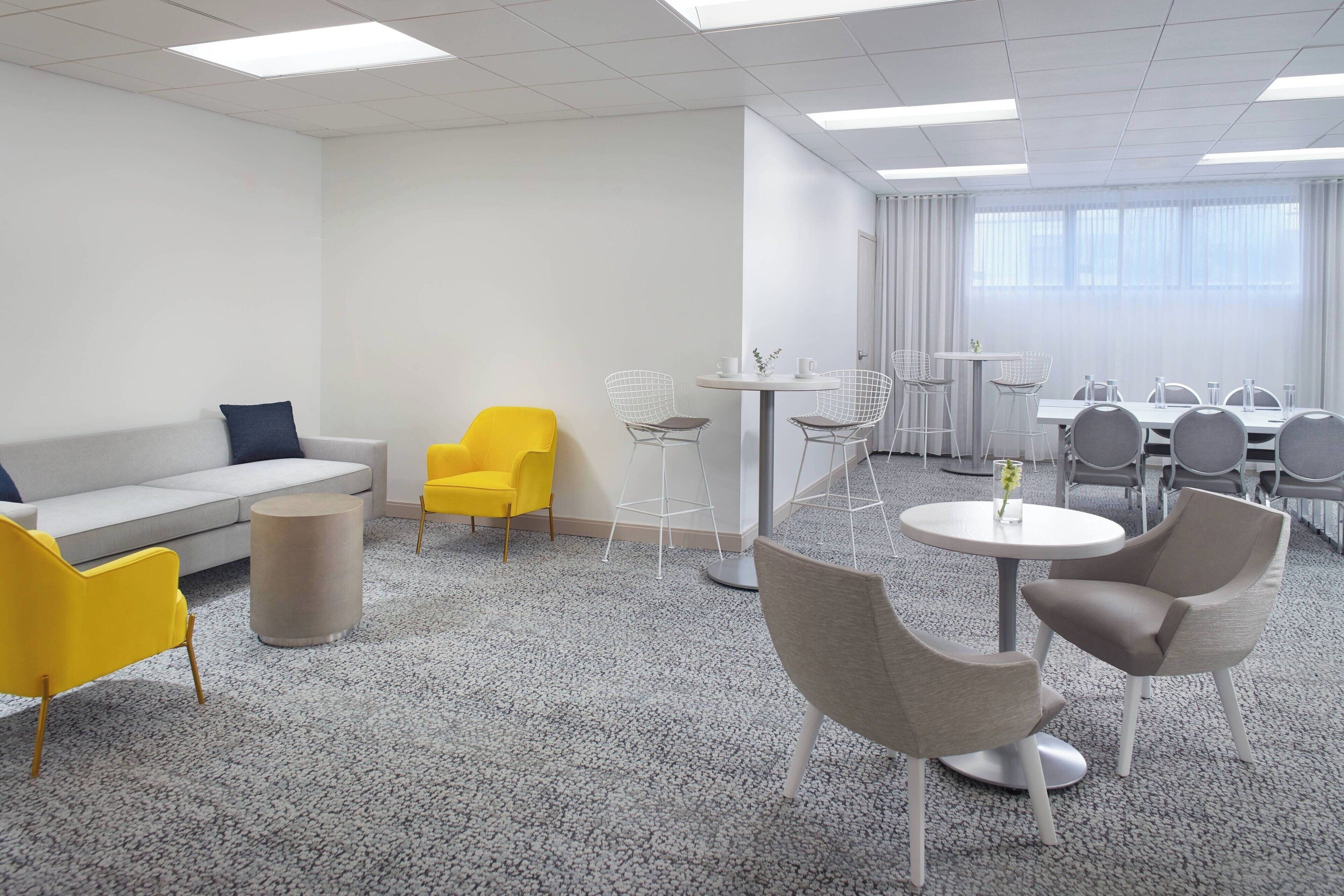 A modern office space with a grey sofa, yellow chairs, a round table with chairs, and a meeting table with chairs.