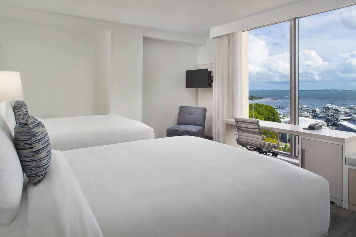 A hotel room with two beds, a desk with a chair, a TV mounted on the wall, and a large window offering a view of the sea and marina.
