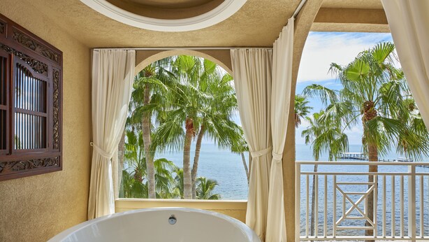 A bathroom with a large bathtub, a window with curtains, and a view of palm trees and the sea. - Cheeca Lodge & Spa (Islamorada)