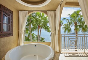 A bathroom with a large bathtub, a window with curtains, and a view of palm trees and the sea.