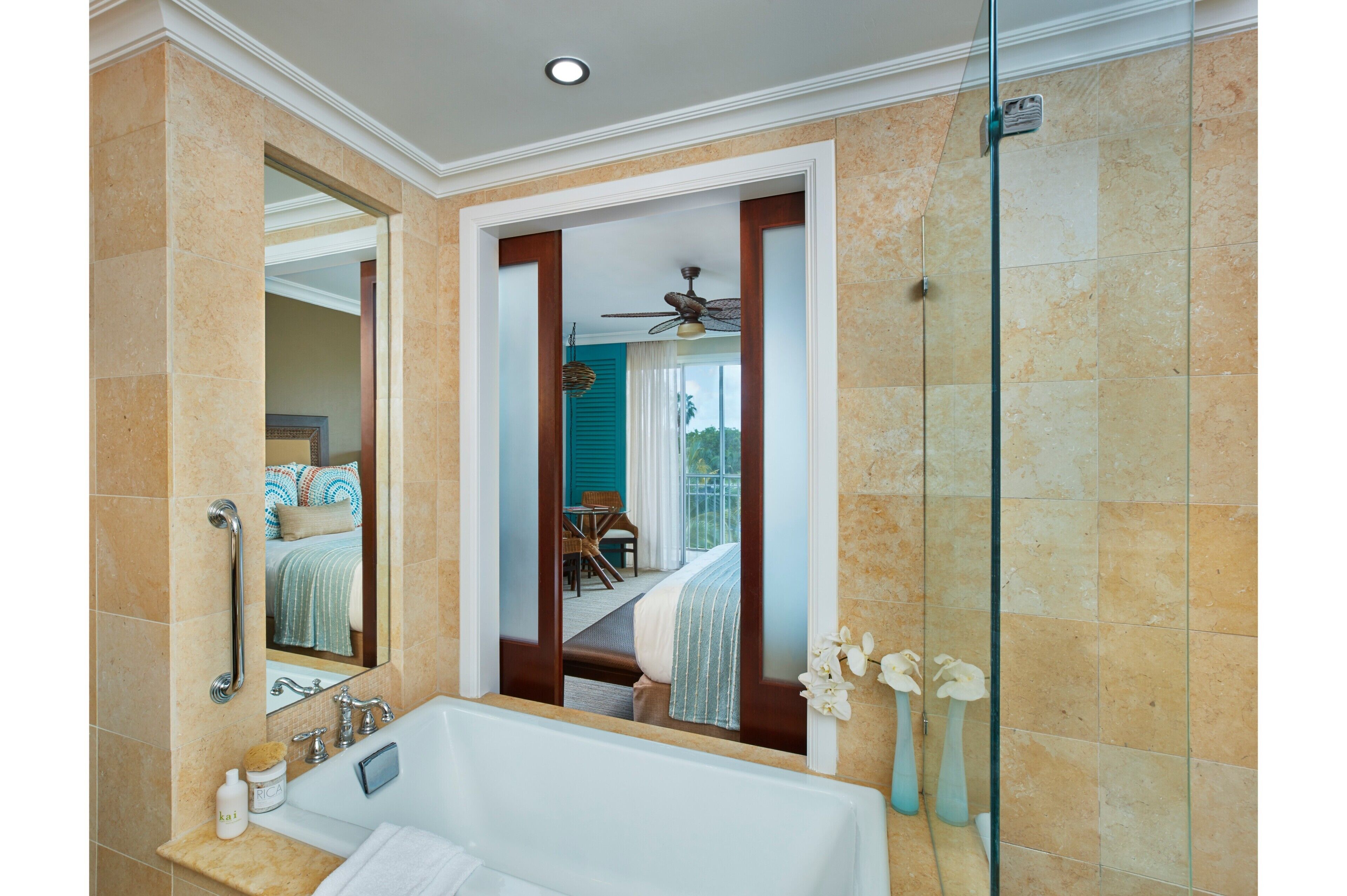 A bathroom with a bathtub, a shower, and a glass door leading to a balcony with a bed and a table.