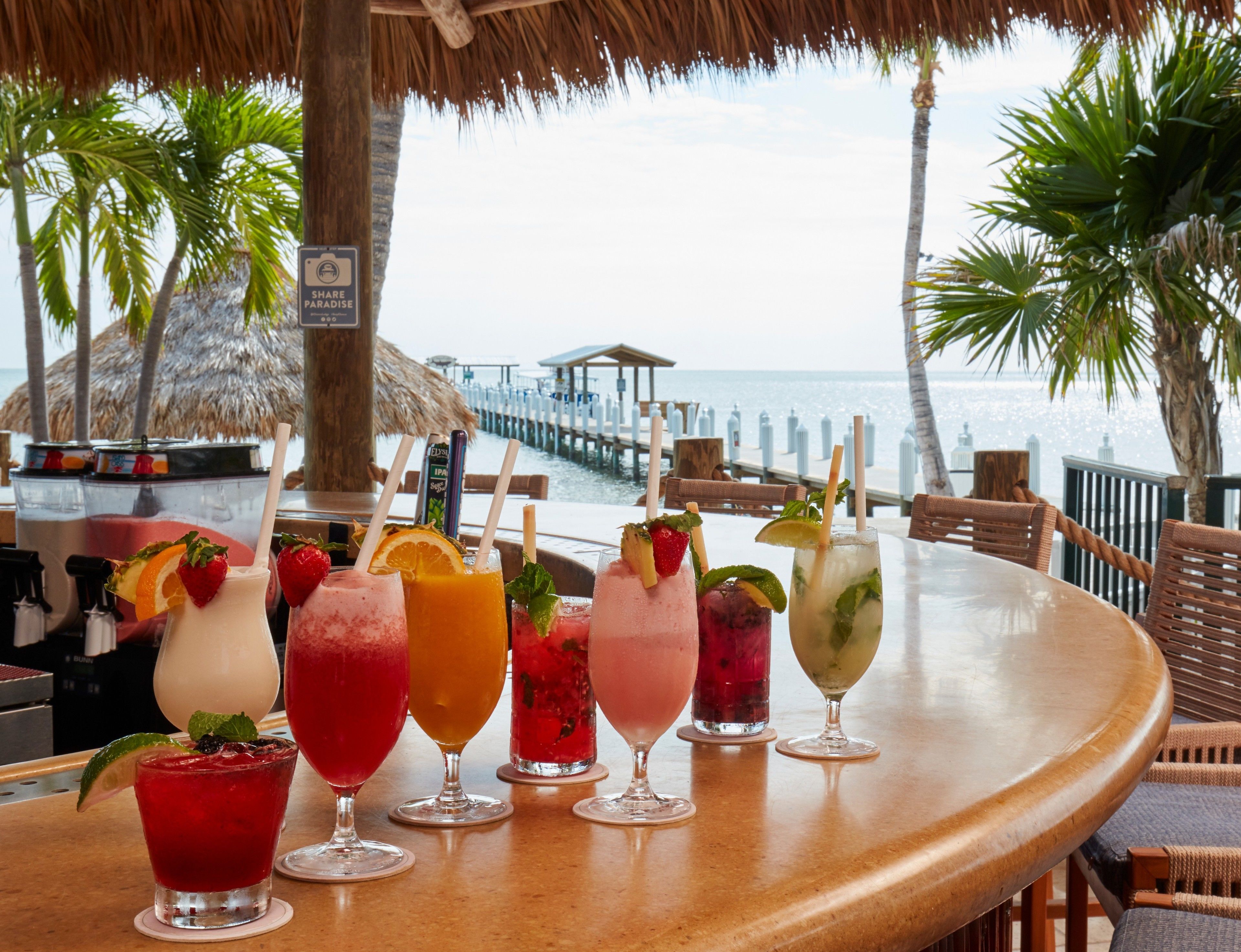 Un bar con diversas bebidas de colores, vista al mar y una estructura de techo de paja.