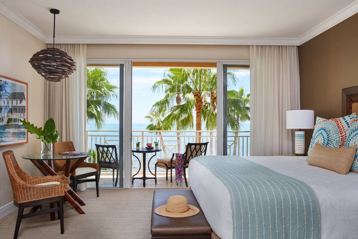 A hotel room with a large bed, a dining table with chairs, a small table with a plant, and a view of palm trees and the ocean.