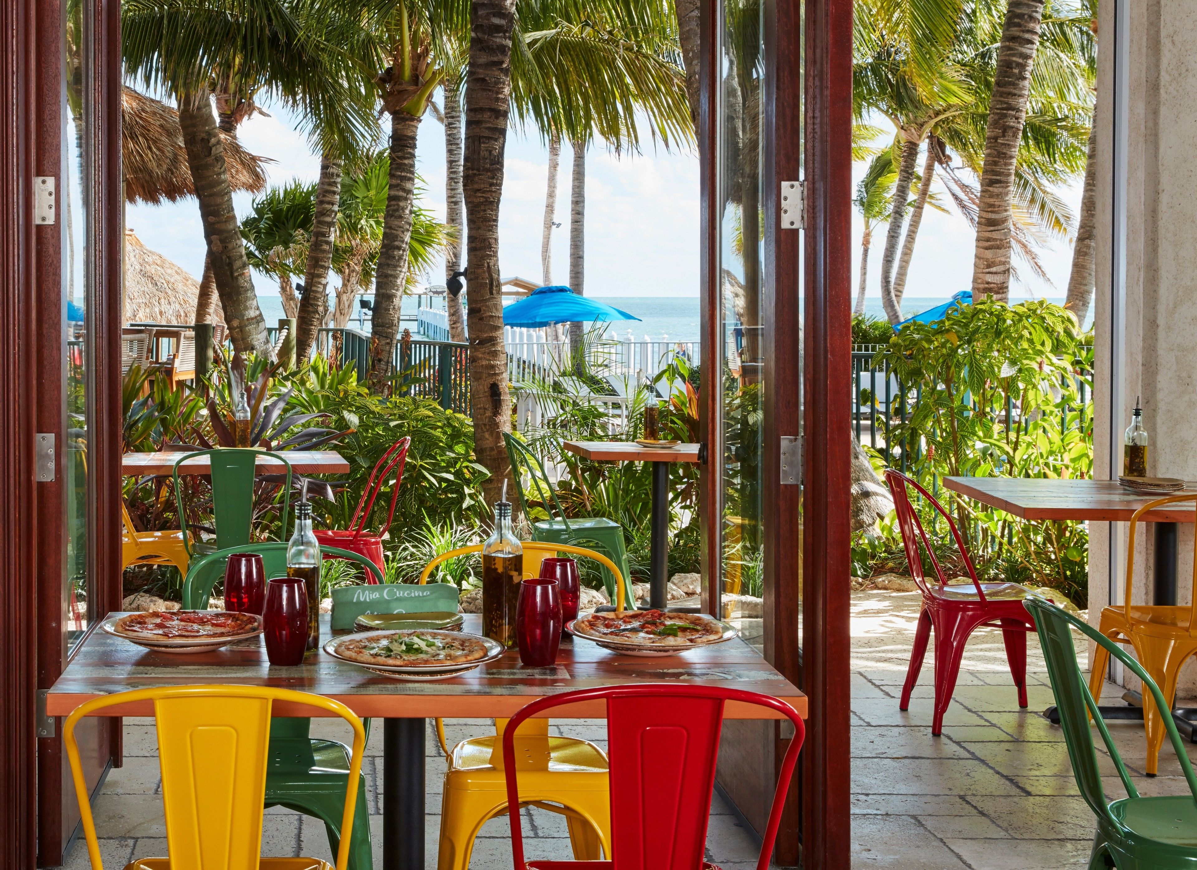An outdoor dining area with colorful chairs and tables, overlooking a beach with palm trees and a thatched-roof structure.