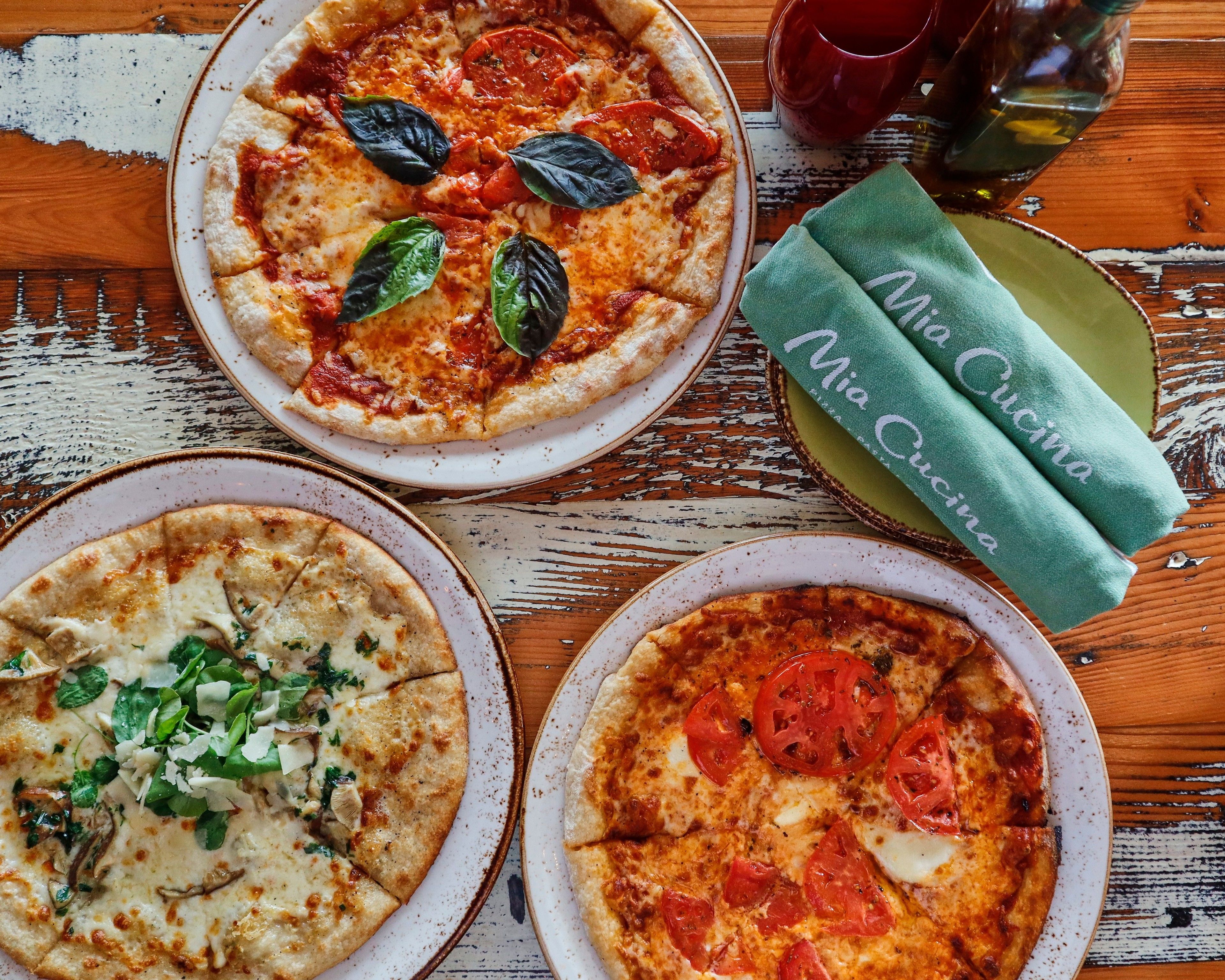 Tres pizzas sobre platos, una mesa de madera rústica y un mantel verde con texto.