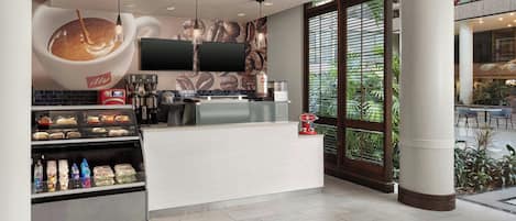 A modern coffee shop with a large coffee cup mural, a counter with a coffee machine, and a display of pastries and bottled drinks.