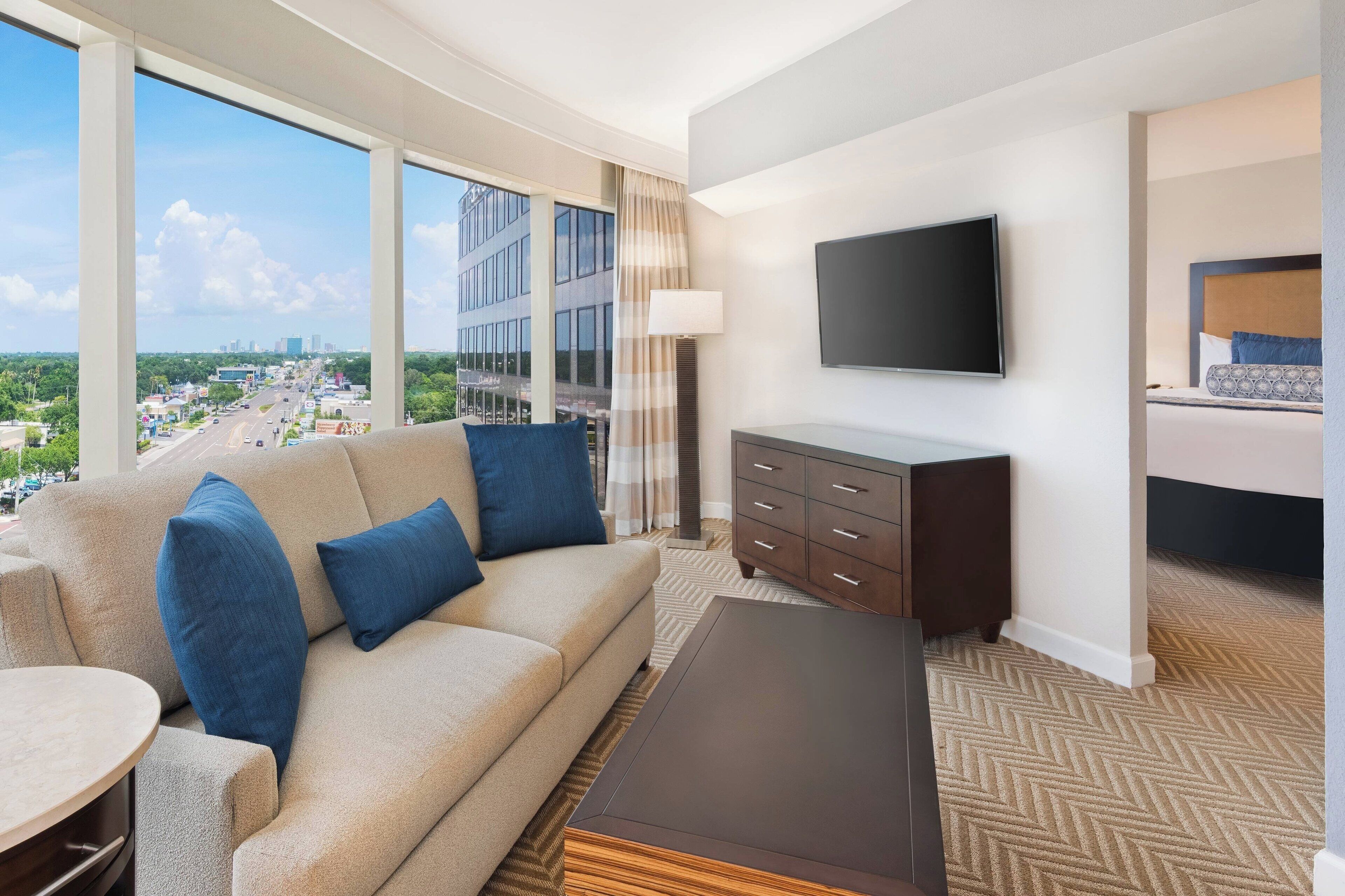 A modern hotel room with a large window offering a city view, a sofa with blue cushions, a flat-screen TV, and a wooden coffee table.
