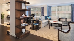 A modern living room with a sectional sofa, a coffee table, and a bookshelf. There is a large window with curtains, a potted plant, and a lamp.