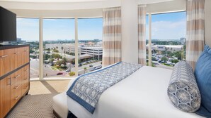 A hotel room with a large bed, a TV, and a view of the city through large windows.