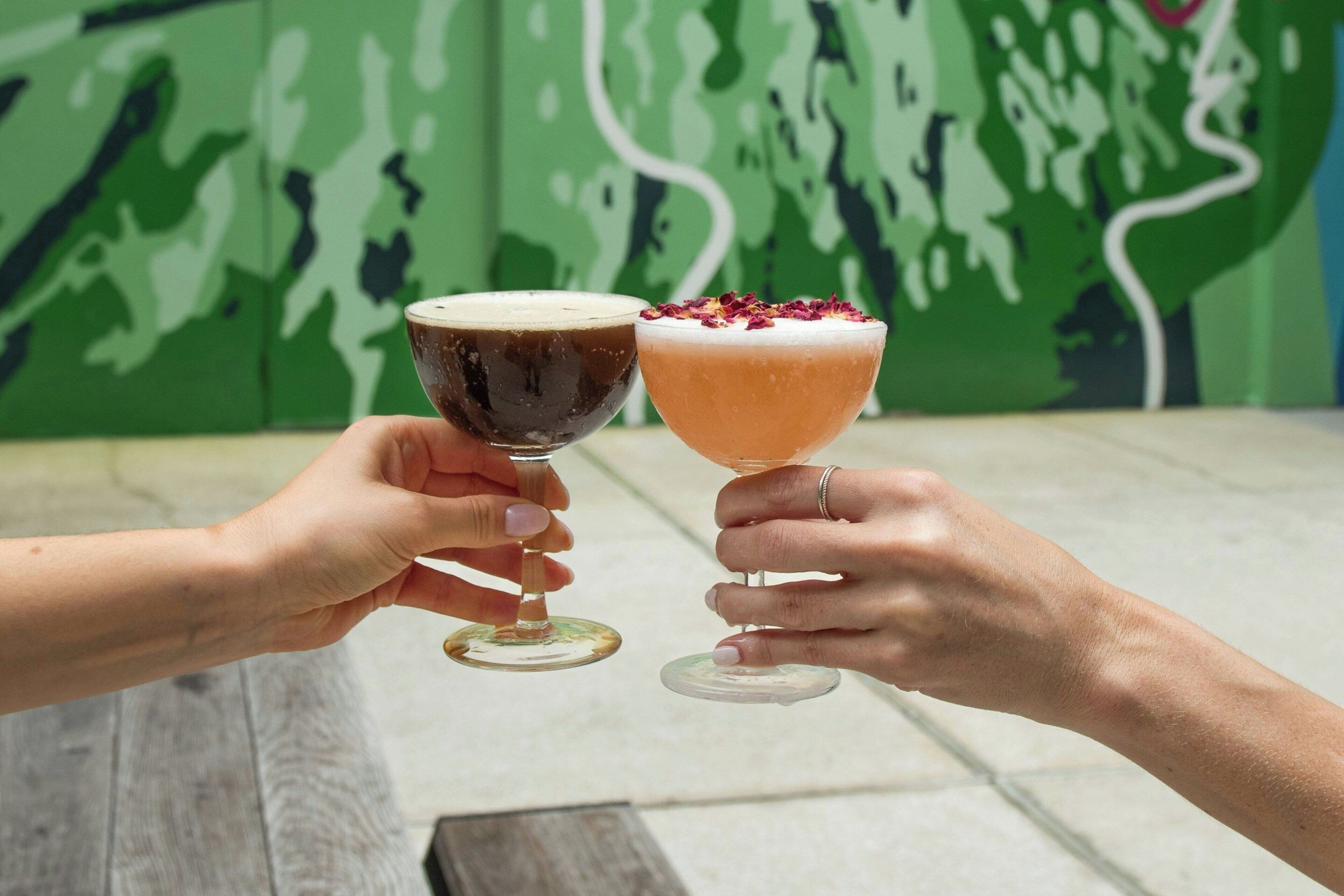 Two hands holding cocktail glasses with a green abstract wall in the background.