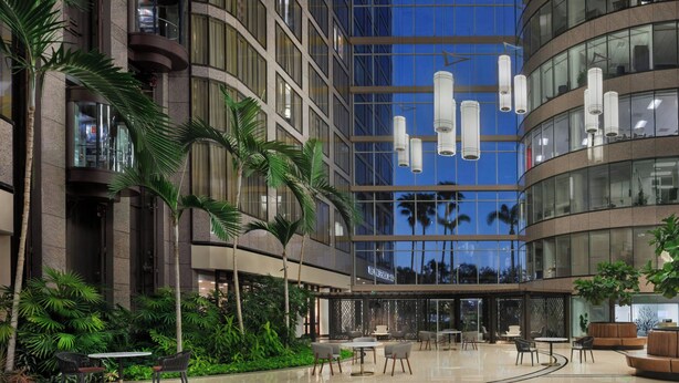 A modern hotel lobby with a glass facade, outdoor seating, and palm trees.