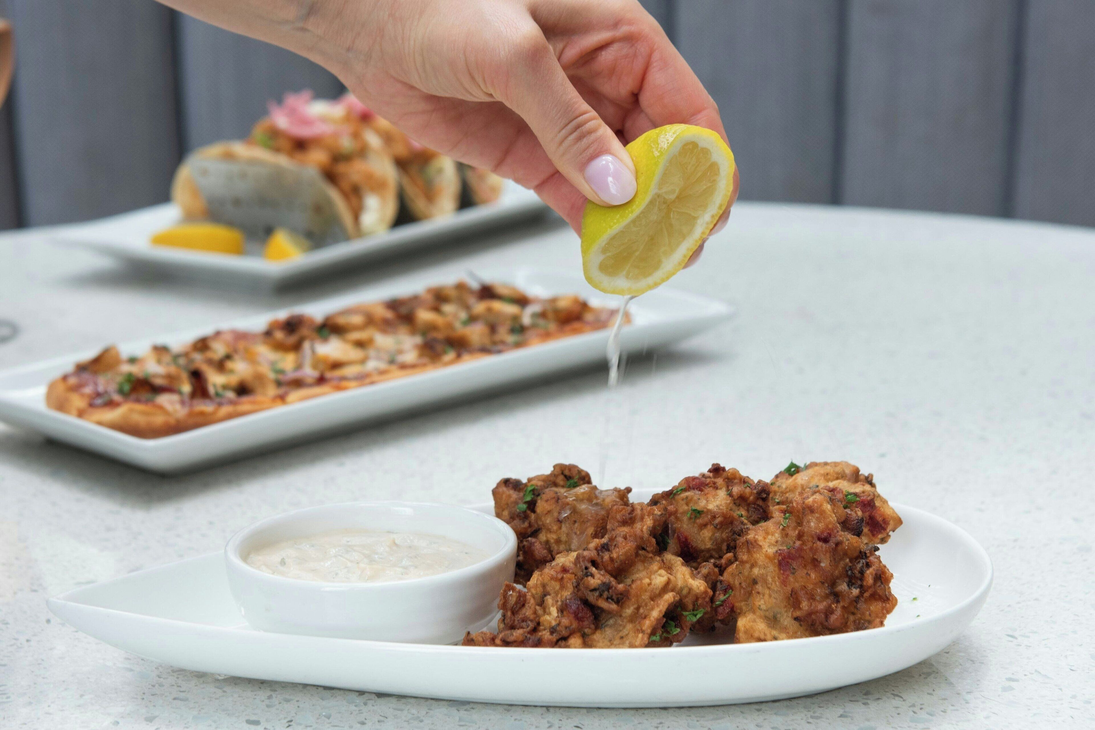 A hand squeezing lemon juice over a plate of fried food, with a bowl of dipping sauce and another plate of food in the background.